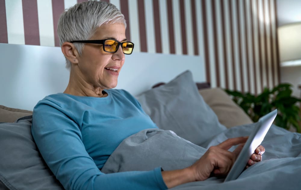 A women in bed using a tablet wearing blue light blocking glasses