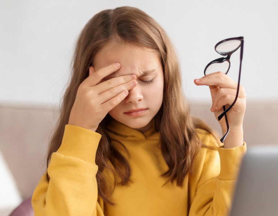 A girl wearing a yellow sweater holding eyeglasses and rubbing her right eye