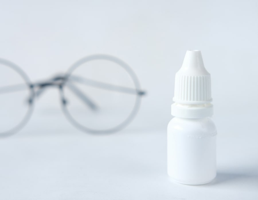 A bottle of eyedrops with a white cap next to a pair of eyeglasses with round frames