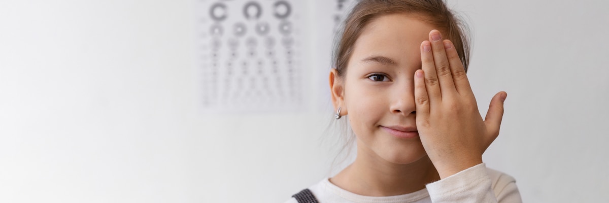 A girl covering one eye with her hand standing in front of an eye chart